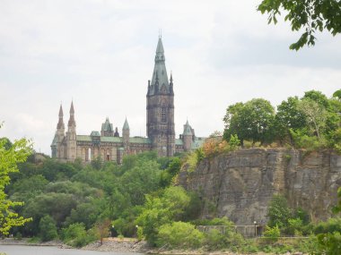 Taş bir uçurumun kenarında bir kale. Bir sürü yeşil ağaç ve açık mavi gökyüzü. Aşağıda bir koşu, bisiklet pisti ve mavi su var. Parliament Hill, Ottawa, Kanada