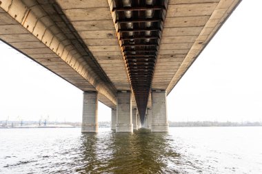 Nehir üzerindeki köprünün gri beton sütunlarının alt manzarası. Köprü iskeleleri, su üzerinde gökyüzü yansıması, perspektif, arkaplan