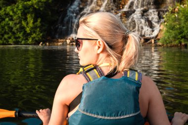 Genç sarışın kadın nehirde kayak yapıyor ve şelale ve ağaçların arka planına bakıyor. Arka plan, yakın plan, portre