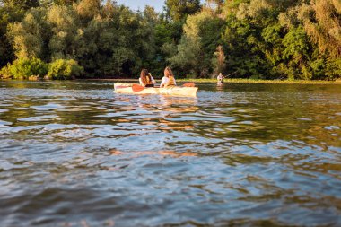Güneşli bir yaz gününde, gün batımında kıyıda balık tutan bir adamın arka planında kanoda fotoğraf çeken iki kız. Hobi konsepti, spor 