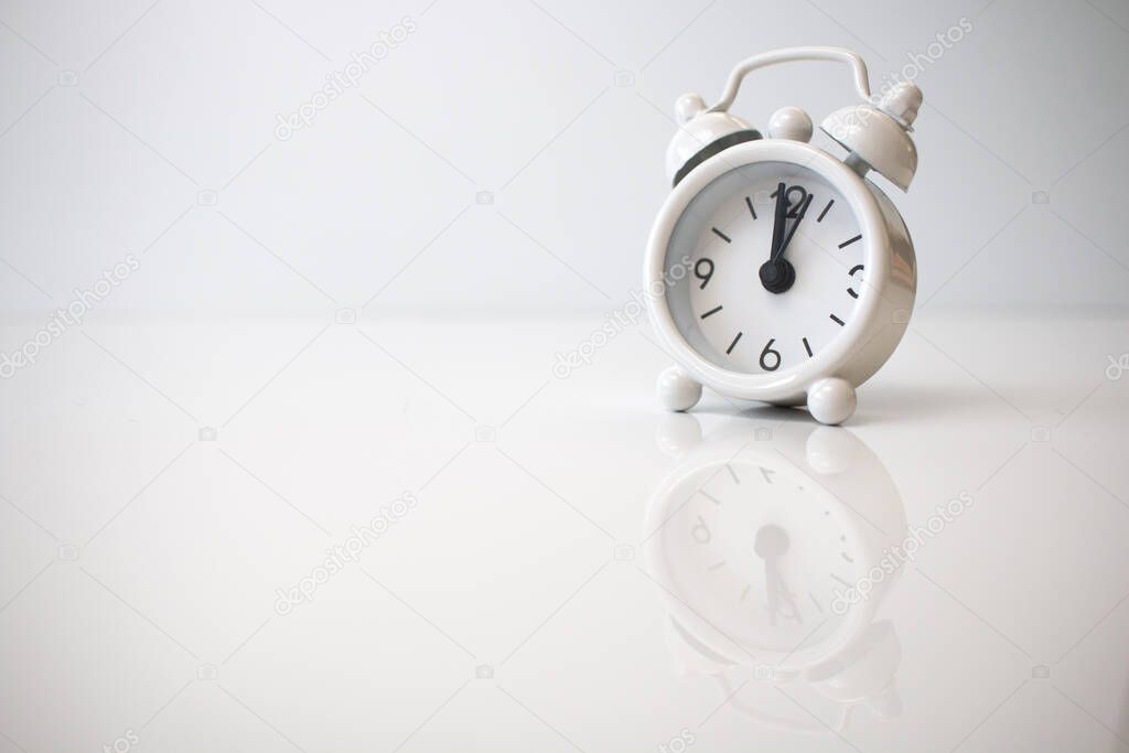 Small analogue white clock showing twelve o'clock with a white background.