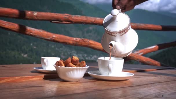 Un tipo sirviendo té de una olla de té blanco en una taza de té con aperitivos mantenidos a lo largo de ella para la hora del té con montañas en el fondo en Manali, Himachal Pradesh — Vídeos de Stock