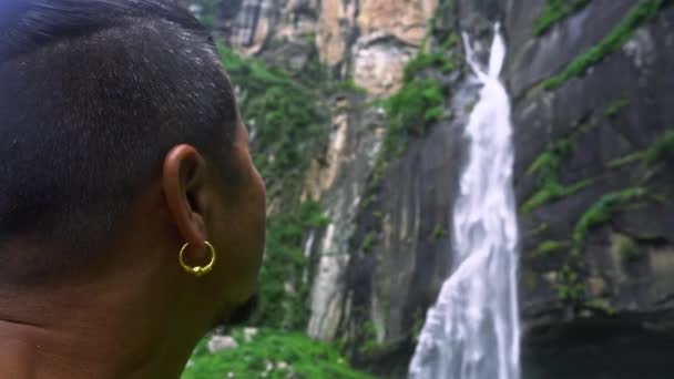 En asiatisk man tittar upp mot ett vattenfall under en molnig dag på Jogini Waterfall, Manali — Stockvideo
