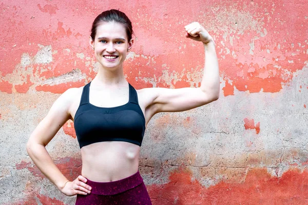 Girl in sport top show arm muscules. Smiling, near red wall copy space