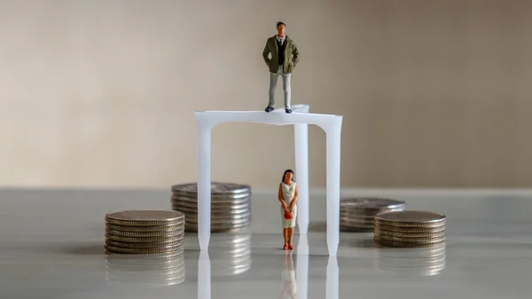 The problem of gender discrimination in pay. A pile of coins beside a miniature man and a miniature woman.
