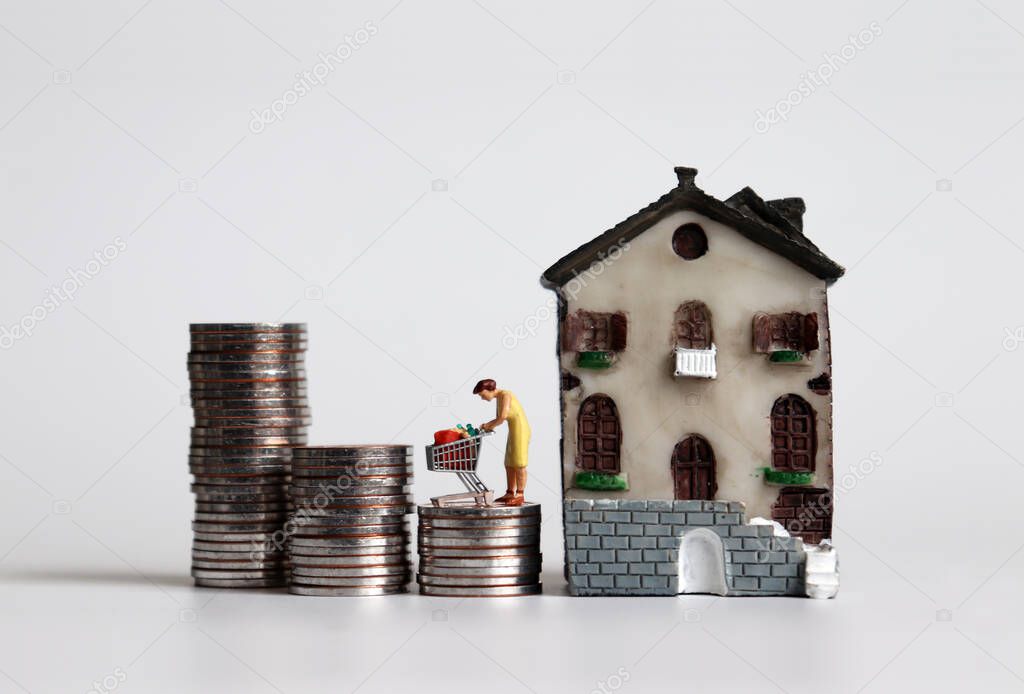 A miniature woman standing with a shopping cart on three pile of stepped coins next to a miniature house.