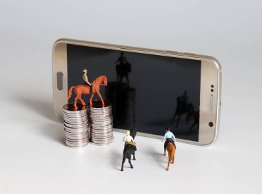 Minyatür bir adam ata biniyor. Akıllı telefon ve bozuk para yığını.