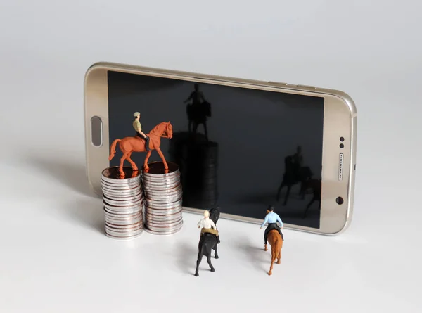 Miniature man riding a horse. Smartphone and pile of coins.