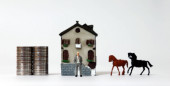 Egy miniatűr ember áll két pénzhalom előtt, egy miniatűr ház és két miniatűr ló..