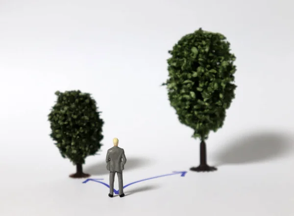 Miniature man with two trees of different sizes on two way. The concept of choice for more profit.