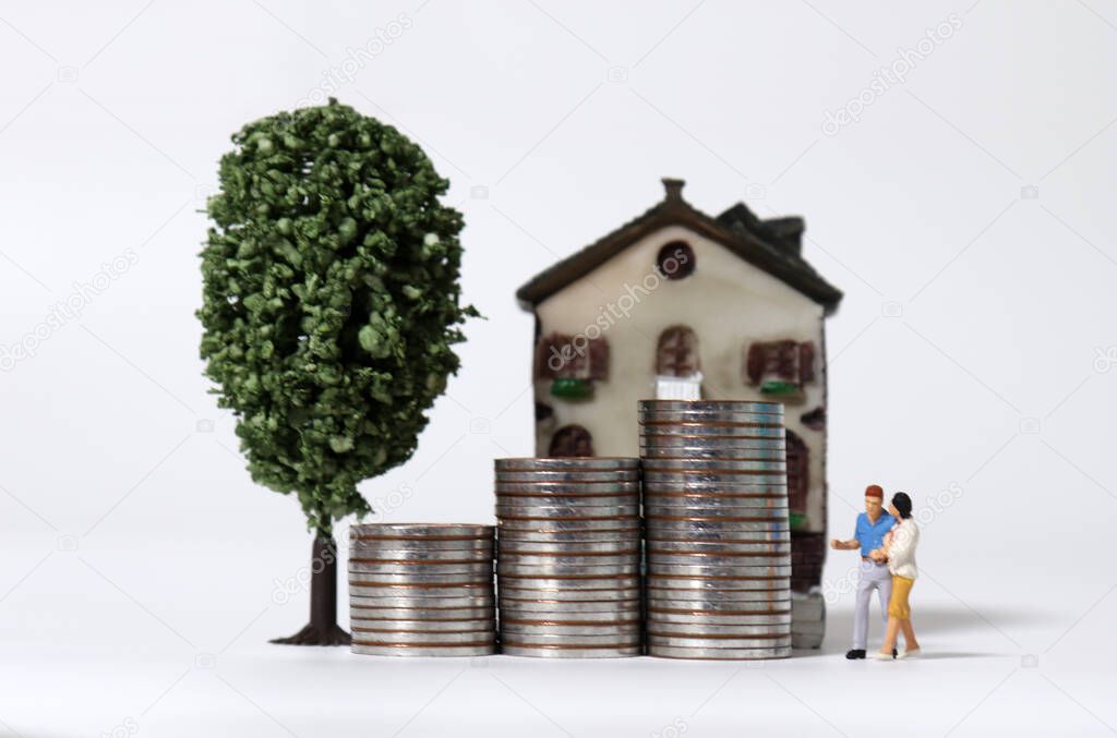 A miniature couple standing with a pile of coins in front of a miniature house.