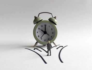 Minyatür adam çalar saatin önünde iki farklı yolda duruyor..