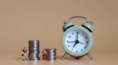 Minyatür bir adamla bozuk para yığınının yanından geçen minyatür bir kadının yanında çalar saat..