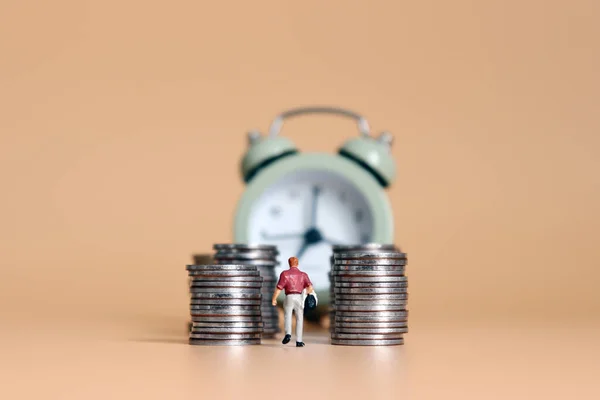A miniature man and an alarm clock walking between piles of coins.