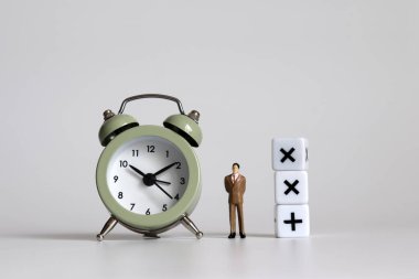 Çalar saat ile matematik operasyon sembolü küpü arasında duran minyatür bir işadamı..