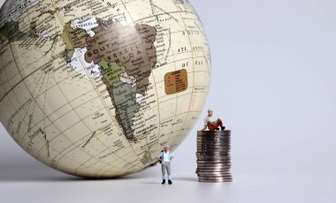 Dünyanın önünde madeni para yığınları olan minyatür turistler. Seyahat maliyeti karşılaştırma kavramı.