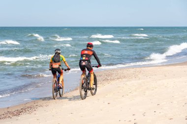 Bisikletli bisikletçiler, arka planda dalgalı Baltık Denizi olan güzel kumlu bir sahilde bisiklet sürüyorlar.
