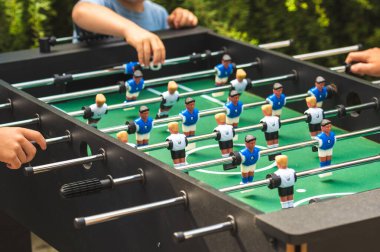 Langırt masasının birkaç oyuncunun ellerinin sapında olduğu yakın plan bir oyun. Masada, karşı takımları temsil eden mavi ve beyaz formalı klasik plastik futbol figürleri yer alıyor.