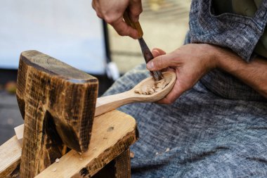Geleneksel ahşap işçiliğini kullanarak tahta kaşığı elle oyan bir zanaatkarın yakın çekimi. Önlüklü bir zanaatkar ahşap talaşı ile çalışıyor. El işçiliği kavramı, el yapımı.