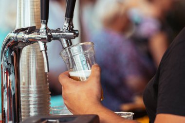 Açık hava festivalinde plastik bardağa taze bira döken bir barmenin yakın çekimi. Musluktan servis edilen soğuk köpüklü bira, ferahlatıcı, demleyici ve kutlamayı sembolize ediyor.