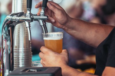Açık hava festivalinde plastik bardağa taze bira döken bir barmenin yakın çekimi. Musluktan servis edilen soğuk köpüklü bira, ferahlatıcı, demleyici ve kutlamayı sembolize ediyor.
