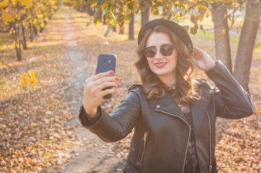 Sonbahar parkında selfie çeken genç bir kadın. Şapkalı, güneşli ve deri ceketli güzel bir kız akıllı telefonuyla kendi fotoğrafını çekiyor..