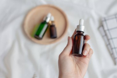 Beyaz kumaş arka planlı bulanık serum şişesinde yüz kremi ya da serum ambalajı tutan kadın elleri. Cilt bakımı için güzellik kozmetik ürünü.