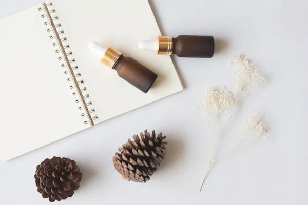 Flat lay of organic aromatherapy oils or skincare products for mock up in minimal style. Dropper glass bottle with notebook, pine cone and dry flower on white background.