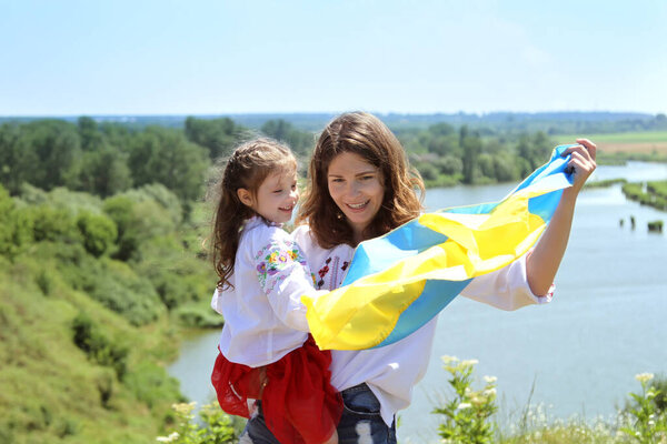 Счастливые улыбающиеся украинская мама и дочь на вышиванке с желто-голубым флагом на фоне гор и реки. День Государственного флага Украины