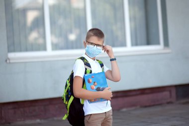 Okula dönelim. Çocuk maske ve sırt çantası takıyor. Koronavirüsten korunuyor. Çocuk salgın hastalık üstüne okula gidiyor. Okulun yanında duruyor. Öğrenciler yeni yıla hazır..