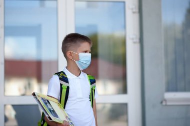 Okula dönelim. Çocuk maske ve sırt çantası takıyor. Koronavirüsten korunuyor. Çocuk salgın hastalık üstüne okula gidiyor. Okulun yanında duruyor. Öğrenciler yeni yıla hazır..