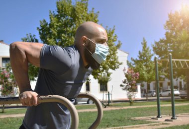 Spor salonundaki adam barda triseps egzersizi yapıyor ve halka açık bir parkta maske takıyor.