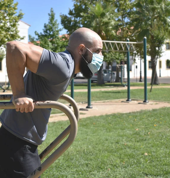 Spor salonundaki adam barda triseps egzersizi yapıyor ve halka açık bir parkta maske takıyor.
