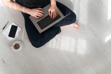 Evde dizüstü bilgisayarı, defteri, kalemi ve kahvesiyle yerde oturan bir adam. İnternetteki ofis. Evden çalışıyorum. Uzayı kopyala Yatay.