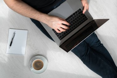 Yerde dizüstü bilgisayarı, defteri, kalemi ve kahvesiyle oturan kot pantolonlu bir adam. Evden çalışıyorum. Bağımsız.