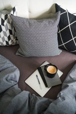 Gri defterde siyah kahve, gümüş dizüstü bilgisayarda, gri battaniyeli, siyah beyaz yastıklı bir kalem. Kahve molası. Evden çalışıyorum. Uzak çalışma. 