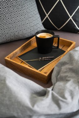 Defterde siyah kahve, ahşap tepside lavanta çiçekleri, gri battaniye ve siyah beyaz yastıklar. Sabah ayini. Yatakta kahve..