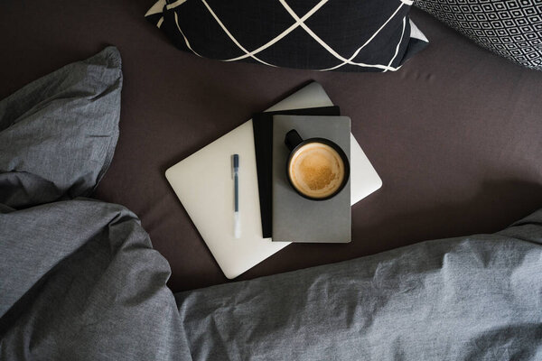 Черная чашка кофе на сером блокноте с ручкой на серебряном ноутбуке на кровати с серым одеялом и черно-белыми подушками. Перерыв на кофе. Работа из дома. Удаленная работа. Вид сверху
.
