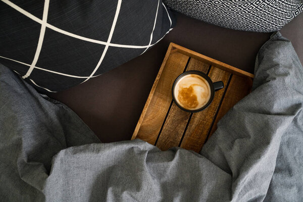 Черная чашка кофе на черном блокноте на деревянном подносе на кровати с серым одеялом и черно-белыми подушками. Утренний ритуал. Вид сверху
.