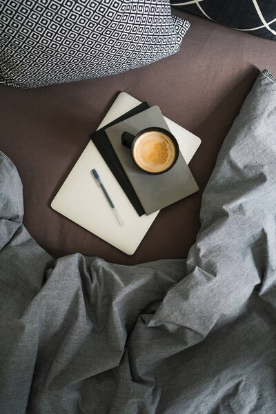 Черная чашка кофе на сером блокноте с ручкой на серебряном ноутбуке на кровати с серым одеялом и черно-белыми подушками. Перерыв на кофе. Работа из дома. Удаленная работа. Вид сверху
.
