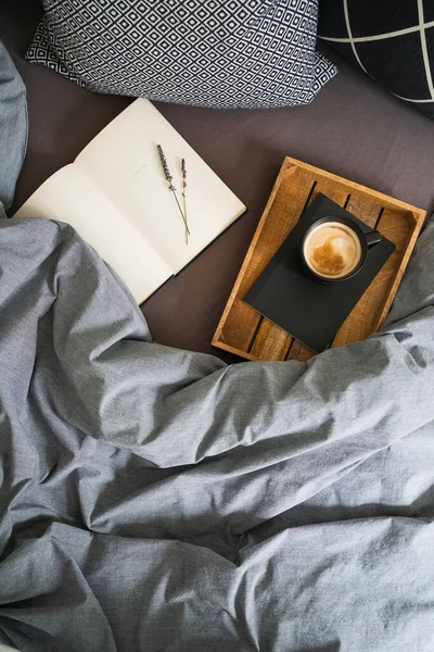 Black cup of coffee on notebook on wooden tray and open book with lavender flowers on the bed with grey blanket and black and white pillows. Morning ritual. Breakfast in bed.