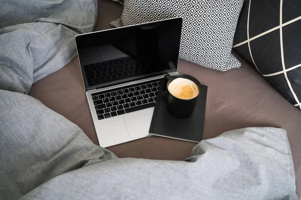 Laptop with notebook and cup of coffee on bed with grey blanket and pillows.