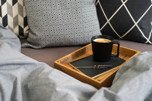 Black cup of coffee on notebook with lavender flowers on wooden tray on bed with grey blanket and black and white pillows. Morning ritual. Breakfast in bed.