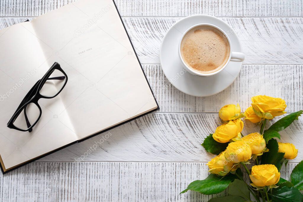 Cup of coffee, yellow peony roses and open book with glasses on white wooden table. Time for break. Top view.