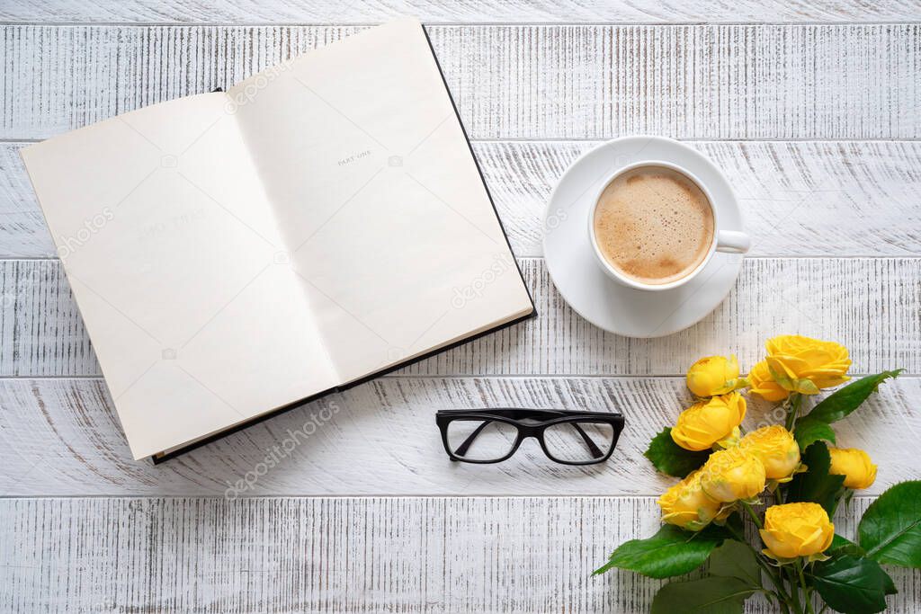 Cup of coffee, yellow peony roses and open book with glasses on white wooden table. Top view. Flat lay.