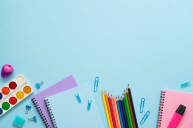 Eğitim konsepti. Mavi arka planda renkli kırtasiye seti. Suluboya, kağıt raptiyeler, mavi, pembe ve mor defterler, silgi, ataç, kalem ve renkli kalemler. Boşluğu kopyala.