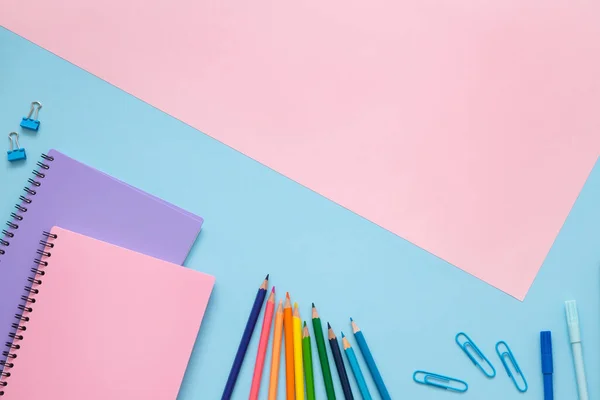 School concept. Purple and pink notebooks, color pencils, felt tip pens and paper clips on blue background and copy space on pink background. Top view.