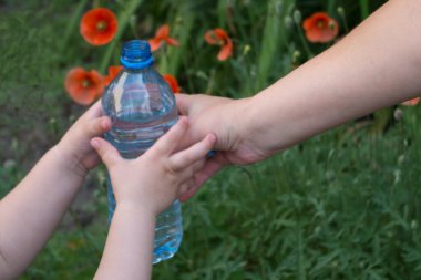 Çocuğun elleri, annenin sağ elinden kırmızı gelincik çiçekleri ve yeşil otlardan oluşan bir arka plan üzerine açık bir şişe su alır. Kavram: çocukluk ve çocuk bakımı