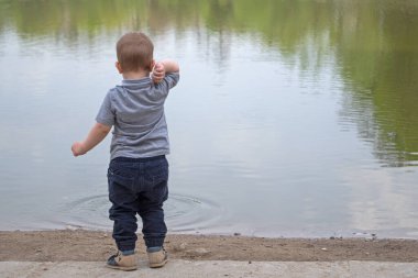 Gri tişörtlü ve kot pantolonlu küçük bir çocuk gölete çakıl taşları atıyor sağ elini sallıyor sahildeki kaldırımda duruyor, suda önceki çakıl taşından dalgalanmaları görebilirsiniz.