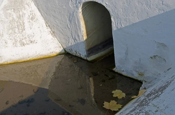 Concrete drainage ditch in the form of an arch whitewashed or painted in white with water after rain and autumn yellow maple leaves under the road or rail surface